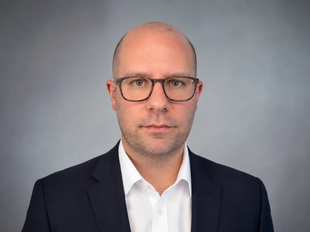 Professional portrait of a bald man wearing glasses, a white shirt, and a dark suit jacket.